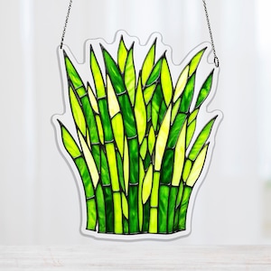 Puede incluir: Panel de vidriera con diseño de planta de bambú. La obra de arte muestra una variedad de tonos verdes y amarillos, con contornos negros que definen los tallos y hojas de bambú. Incluye una cadena para colgar.