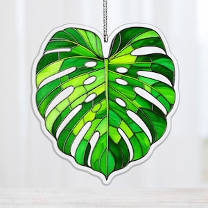 May include: A stained glass suncatcher shaped like a Monstera leaf. The leaf features various shades of green glass, with white lines dividing the sections. A silver chain is attached for hanging.