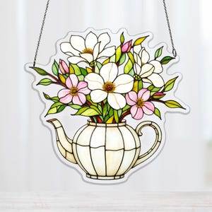 以下が含まれることがあります： 白とピンクのモクレンの花と緑の葉で満たされた白いティーポットを描いたステンドグラスの窓パネル。 パネルは黒で縁取られ、チェーンから吊り下げられています。