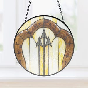 May include: A round stained glass window with a black metal frame and chain for hanging. The design features a central motif of three silver arrows against a white and yellow background, framed by a brown, wood-grain-patterned arch.