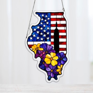 Op de afbeelding: Gebrandschilderd glaskunstwerk in de vorm van Illinois, met de Amerikaanse vlag, een silhouet van een wolkenkrabber en kleurrijke bloemen. Het heeft een witte rand en hangt aan een zilveren ketting. Ontworpen om op te hangen.