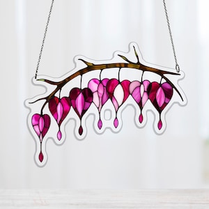 Pode incluir: Obra de arte em vitral com um galho com várias flores em forma de coração em tons de rosa e magenta. As flores têm um efeito de gradiente, com bordas mais escuras e centros mais claros. A peça é suspensa por uma corrente.
