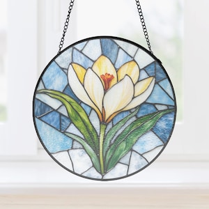 Puede incluir: Vidriera redonda con una flor de crocus amarilla y blanca con hojas verdes. La flor está centrada en un fondo de mosaico azul y blanco, enmarcada por un borde negro y una cadena para colgar.