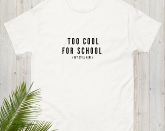 May include: White t-shirt with the text "TOO COOL FOR SCHOOL (BUT STILL HERE)" in black, centered on the front. The shirt is made of a soft material and has short sleeves. The background is a light wood surface.