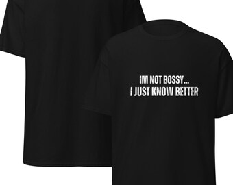 以下が含まれることがあります： 「IM NOT BOSSY... I JUST KNOW BETTER」の白文字がプリントされた黒いTシャツ。画像には、同じデザインのTシャツが2枚写っており、1枚は手前に、もう1枚は背景にあります。