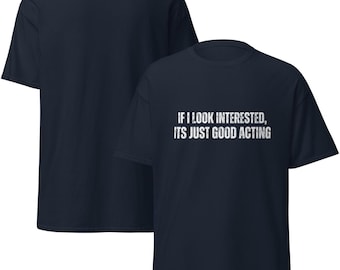 Op de afbeelding: Marineblauw T-shirt met de witte tekst "IF I LOOK INTERESTED, ITS JUST GOOD ACTING". Het T-shirt heeft korte mouwen en een ronde hals. Een ander marineblauw T-shirt is gedeeltelijk zichtbaar op de achtergrond.