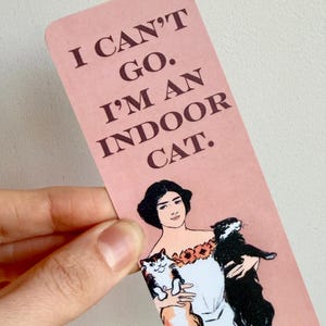 May include: A pink bookmark with the text "I CAN'T GO. I'M AN INDOOR CAT." features an illustration of a woman holding two cats. The woman is wearing a white dress and a floral crown. One cat is black and white, the other is white and orange.