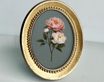 May include: An oval-shaped picture frame with a metallic gold finish and a beaded inner border. The frame contains a print of two pink peony blossoms and a small bud with green leaves set against a solid, muted gray-blue background.