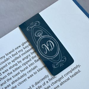 May include: A navy blue bookmark with a white decorative design and the initials "ND" inside a circular frame. The bookmark is placed on a book with printed text.