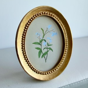 May include: A gold-colored, oval picture frame with a beaded border. The frame holds a botanical print of blue flowers and green leaves against a light beige background. The frame is standing upright on a white surface.