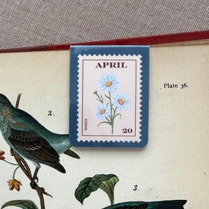 May include: A blue and white floral stamp-shaped bookmark with the word "APRIL" at the top and the number "20" at the bottom. The bookmark features a light blue flower design. The bookmark is placed on a vintage book page with illustrations of birds.