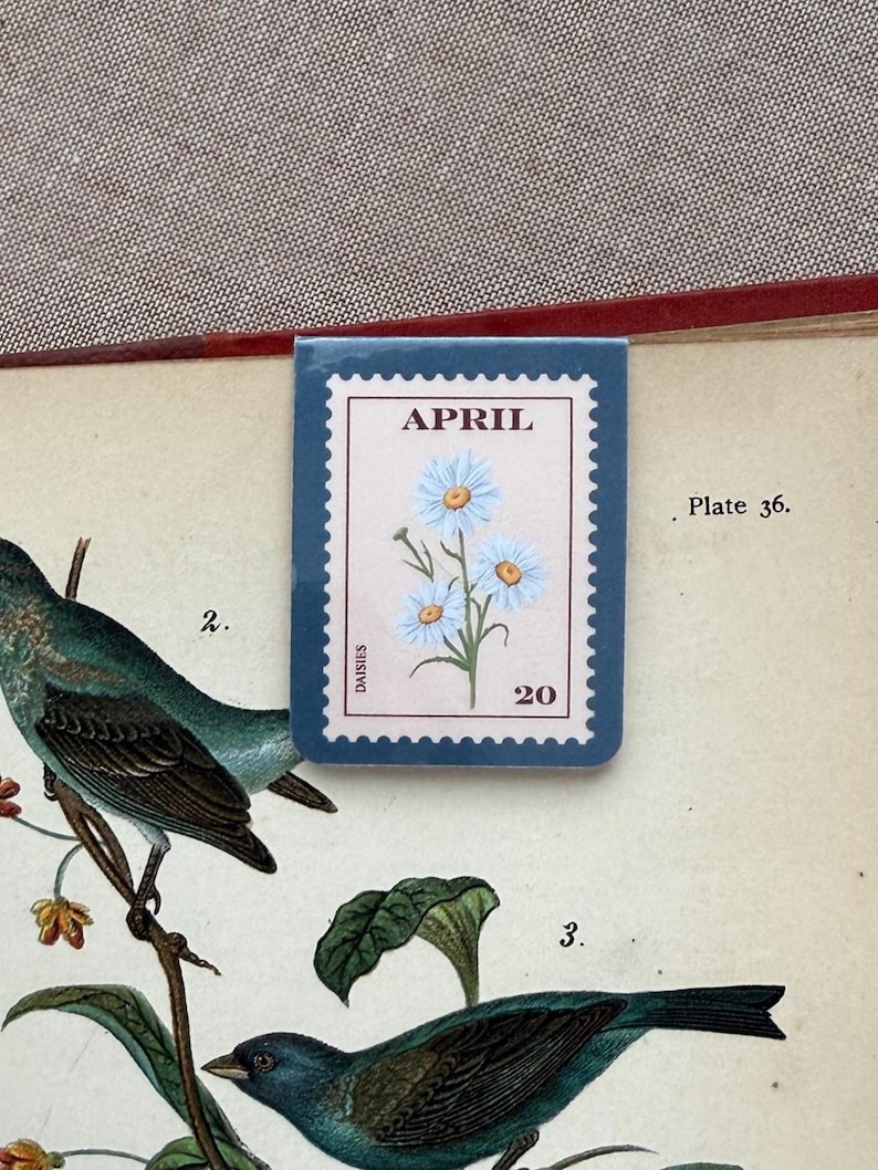 May include: A blue and white floral stamp-shaped bookmark with the word "APRIL" at the top and the number "20" at the bottom. The bookmark features a light blue flower design. The bookmark is placed on a vintage book page with illustrations of birds.