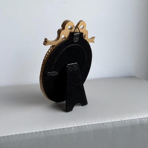 May include: A small, gold-colored oval picture frame with a decorative bow at the top. The frame has a black backing and a black stand. The frame is on a white and gray surface.