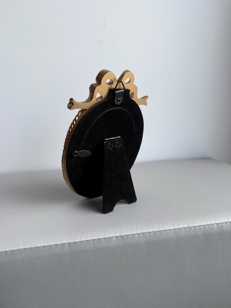 May include: A small, gold-colored oval picture frame with a decorative bow at the top. The frame has a black backing and a black stand. The frame is on a white and gray surface.