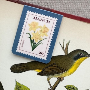 May include: A blue bookmark with a stamp design featuring yellow daffodils and the text "MARCH" and "20". The bookmark is placed over a botanical illustration of a bird perched on a branch.