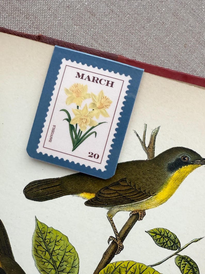 May include: A blue bookmark with a stamp design featuring yellow daffodils and the text "MARCH" and "20". The bookmark is placed over a botanical illustration of a bird perched on a branch.