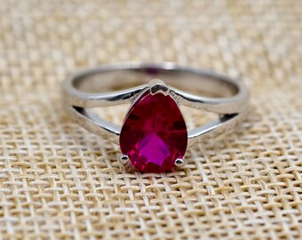 Peut inclure: Bague en argent ornée d'une pierre rouge foncé en forme de poire. L'anneau se divise en deux lignes parallèles de chaque côté de la pierre, créant un design unique. La bague est posée sur un fond beige texturé.