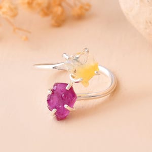May include: A silver ring featuring two raw gemstones. One gemstone is a vibrant magenta color, and the other is a combination of yellow and clear. The ring has a modern, open design.