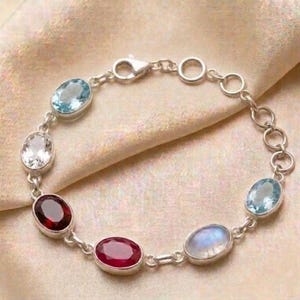 May include: A silver bracelet featuring a series of oval-shaped gemstones in various colors, including blue, red, and clear. The bracelet is displayed on a cream-colored fabric.