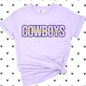 May include: A light purple short-sleeve t-shirt featuring the word COWBOYS in a white and purple gingham pattern with a dark purple outline. The word d'hanis is written in a gold cursive script font layered over the center of the text.