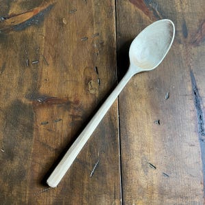 May include: A light-colored wooden spoon with a long handle and oval bowl. The spoon is made of a light-colored wood and has a smooth finish. The spoon is resting on a dark brown wooden surface.