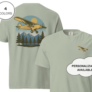 Puede incluir: Camiseta verde salvia con un gráfico de avión amarillo en la espalda, con un fondo de montañas y bosque. Un gráfico de avión más pequeño está en la parte delantera. El texto en la imagen dice "¡Personalización disponible!" y "4 colores".