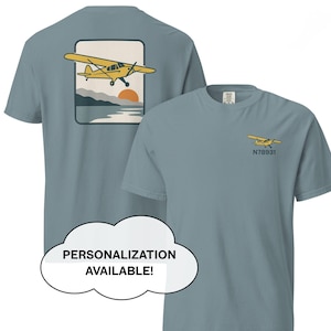 Puede incluir: Dos camisetas azul grisáceo. Una camiseta tiene un pequeño gráfico de avión amarillo con el texto "N78931". La otra camiseta tiene un gráfico más grande de un avión amarillo sobre un paisaje con una puesta de sol. Texto en una burbuja de diálogo: "¡PERSONALIZACIÓN DISPONIBLE!"
