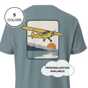Puede incluir: Camiseta azul polvoriento con un gráfico de avión amarillo dentro de un marco rectangular. El gráfico incluye una puesta de sol, montañas y agua. El texto en la camiseta indica que la personalización está disponible y que la camiseta está disponible en 5 colores.