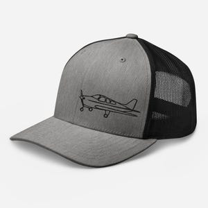 Könnte beinhalten: Graue Trucker-Kappe mit schwarzem Mesh-Rücken. Die Kappe zeigt eine schwarze Umrisszeichnung eines kleinen Flugzeugs auf der Vorderseite. Die Kappe hat einen gebogenen Schirm und einen Snapback-Verschluss.