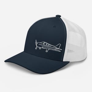 Könnte beinhalten: Marineblaue Trucker-Cap mit weißem Mesh-Rücken und gebogenem Schirm. Auf der Vorderseite der Cap ist ein weißer, gestickter Umriss eines kleinen Flugzeugs zu sehen. Mit Druckknopfverschluss für verstellbare Größe.