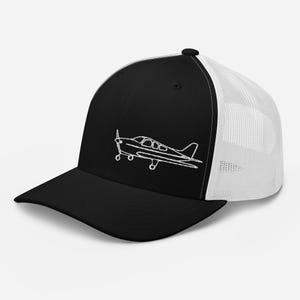 Könnte beinhalten: Schwarz-weiße Trucker-Cap mit weißer, gestickter Umrisszeichnung eines kleinen Flugzeugs auf der Vorderseite. Die Kappe hat eine schwarze Vorderseite und einen Schirm, mit einem weißen Mesh-Rücken und einem gebogenen Schirm.