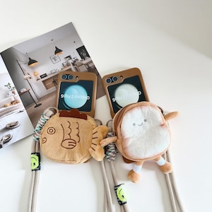 May include: Two brown phone cases for Galaxy Z Flip5 with plush attachments. One is a fish design, the other is a slice of bread with a smiling face. Each case has a beige lanyard with a green smiley face.
