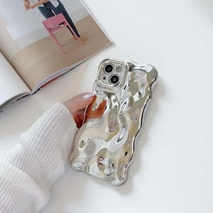 Puede incluir: Funda de teléfono plateada con un diseño ondulado único. La funda tiene una superficie brillante y reflectante y un recorte para la cámara del teléfono. La funda la sostiene una persona con un suéter blanco. Accesorio de teléfono.