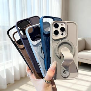 Puede incluir: Una colección de fundas para teléfonos en varios colores: negro, marrón, morado, azul claro y gris. Cada funda tiene un agarre circular y un recorte para la cámara. Las fundas están diseñadas para proteger el teléfono.