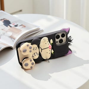 May include: Black phone case with a 3D cat design. The case features a black body with a cream-colored face, paws, and a pink nose. The cat's ears are purple, and the eyes are green. The phone case is lying on a white surface.