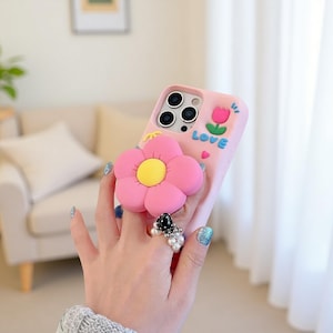 Puede incluir: Funda de teléfono rosa con un diseño de flor en 3D. La flor tiene pétalos rosas y un centro amarillo. La funda también presenta un tulipán y la palabra "LOVE" en letras azules. La funda del teléfono la sostiene una mano con varios anillos.
