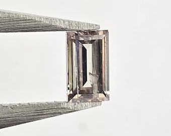May include: A rectangular, light-colored gemstone held by tweezers. The gem has a pale pinkish-brown hue and a rectangular cut with visible facets. The tweezers are silver-colored and hold the gem against a white background.