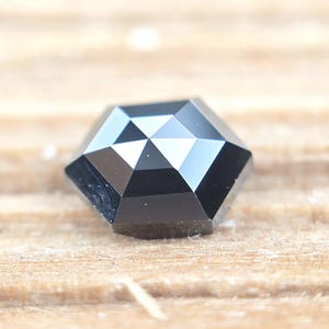 Pode incluir: Uma pedra preciosa preta facetada com formato hexagonal. A pedra apresenta uma superfície superior plana com múltiplas facetas triangulares que irradiam do centro, criando um padrão geométrico com bordas afiadas e um acabamento preto opaco polido.
