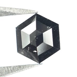Può includere: Un diamante nero sciolto a taglio esagonale con superficie sfaccettata, che mostra un aspetto scuro e opaco con sottili riflessi interni e bordi geometrici affilati, tenuto da pinzette metalliche su uno sfondo bianco.