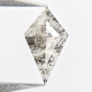 May include: A kite-shaped salt and pepper diamond with a faceted surface, featuring a translucent appearance with dense black and grey inclusions throughout the stone, held securely by two metal tweezers at the top and bottom points.