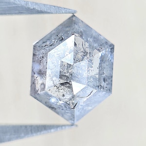 May include: A hexagonal, salt and pepper diamond with a translucent, icy blue-grey hue. The gemstone features a faceted surface with visible internal inclusions, dark carbon spots, and a geometric cut that creates a multi-faceted appearance.