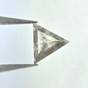 May include: A triangular, rough-cut diamond held by tweezers. The gemstone has a gray and white mottled appearance with a translucent quality. The edges of the diamond are sharp and well-defined, and the overall shape is a perfect equilateral triangle.