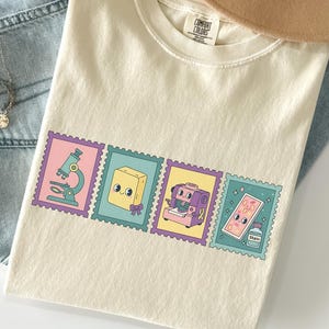 May include: Cream-colored t-shirt with four colorful stamp designs featuring a microscope, a book, a sewing machine, and a stain remover. The shirt is paired with light blue jeans, a tan hat, and a necklace.