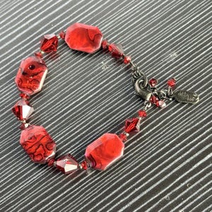 May include: Red beaded bracelet featuring large, faceted, rectangular beads with a marbled effect. The bracelet also includes smaller, faceted, red crystal beads and a silver-tone clasp. A small metal tag is attached.