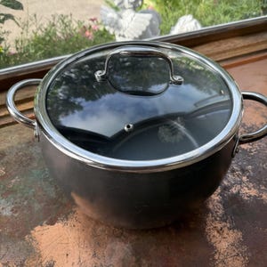 Può includere: Una pentola nera con coperchio in vetro trasparente e bordo argentato. La pentola ha due manici argentati e si trova su una superficie color rame. Il coperchio ha una maniglia argentata.