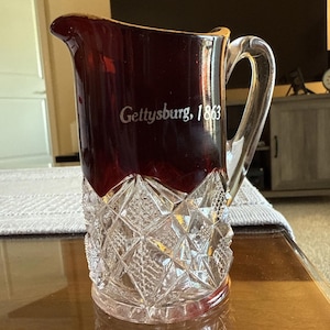 May include: A clear glass pitcher with a ruby red top and a diamond-cut base. The pitcher has a clear handle and the words "Gettysburg, 1863" are printed on the side. The pitcher is sitting on a wooden surface.
