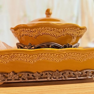 May include: Mustard yellow ceramic serving dish with a lid, set on a decorative metal stand, and placed on a matching rectangular tray. The dish and tray feature ornate, embossed scrollwork and floral accents, creating an elegant presentation.