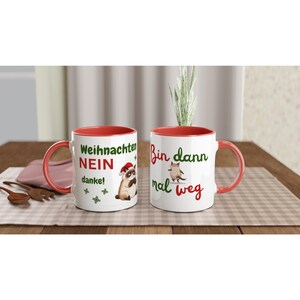 Könnte beinhalten: Zwei weiße Keramikbecher mit roten Innenräumen und Griffen. Ein Becher zeigt einen Waschbären mit Weihnachtsmütze und dem Text "Weihnachten NEIN danke!". Der andere Becher hat eine Katze und den Text "Bin dann mal weg".