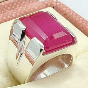 May include: A silver ring with a large, rectangular, magenta-colored gemstone. The ring has a wide band and a modern design, set against a cream-colored surface within a pink dotted box.