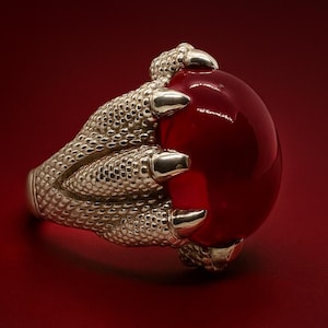 Pode incluir: Anel prateado com um design detalhado de garra segurando uma grande pedra preciosa vermelha polida. O anel tem uma superfície texturizada e está sobre um fundo vermelho escuro.